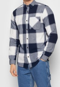 Camisa de cuadros de manga larga en colores azul, marino y blanco con cuello abotonado y bolsillo en el pecho. Hecha de tela texturizada.