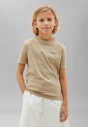 Blond dziecko o włosach sięgających do ramion nosi beżowy t-shirt Calvin Klein i białe szorty, stoi na tle jednolitego szarego tła.