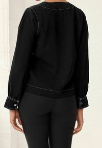 Schwarze Bluse mit langen, gepufften Ärmeln und weißen Kontrastnähten. Verfügt über einen abgerundeten Ausschnitt und Manschettendetails, kombiniert mit eng anliegenden schwarzen Hosen.