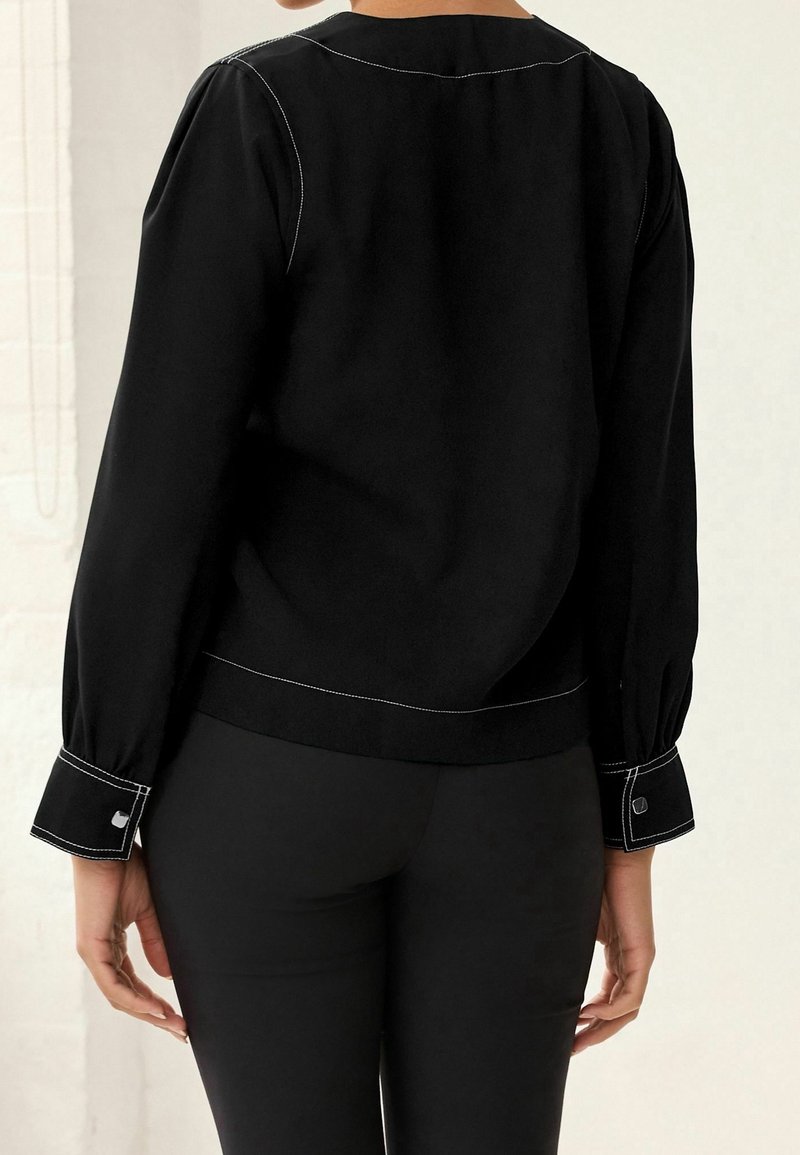 Schwarze Bluse mit langen, gepufften Ärmeln und weißen Kontrastnähten. Verfügt über einen abgerundeten Ausschnitt und Manschettendetails, kombiniert mit eng anliegenden schwarzen Hosen.