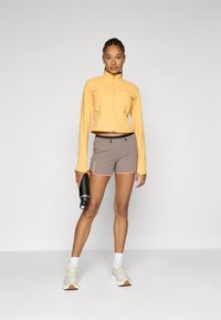Lätt, gul långärmad träningströja, beige shorts med rosa kant, och vita sneakers. Modell som håller en svart vattenflaska.