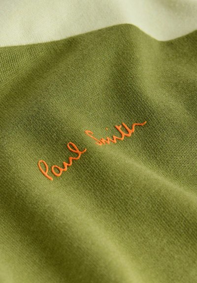Grønt stof med orange broderet tekst i signaturstil, hvor der står "Paul Smith" på en let krøllet overflade.