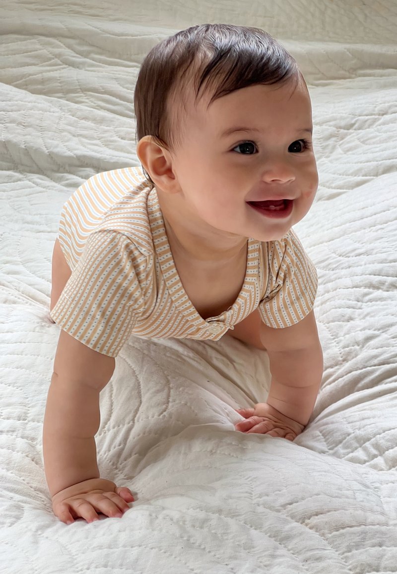 Kortermet body med lyse oransje og hvite striper, med trykknappfeste og myk, teksturert stoff. Babyen kryper på et quiltet teppe.