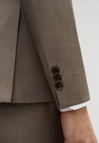 Rękaw brązowej marynarki garniturowej z trzema ciemnymi guzikami i beżowym przeszyciem, ukazujący biały mankiet koszuli pod spodem na ramieniu osoby.