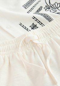 Ceinture blanche élastique avec cordon sur tissu texturé, couvrant partiellement un vêtement blanc avec des motifs géométriques et floraux noirs.