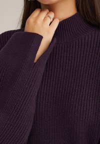Femme portant un pull en maille ribbed violet foncé avec un col haut, touchant son menton avec sa main gauche montrant une bague en argent à son doigt.