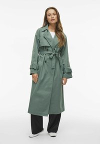 Cappotto trench verde lungo con vita regolabile, bottoni doppi, ampi rever e polsini regolabili; texture liscia, materiale leggero.