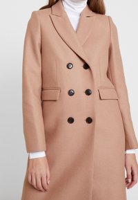 Dobbeltbrystet trenchcoat i lysebrunt stof, med sorte knapper, slagkraver og sidelommer. Glat, skræddersyet design.