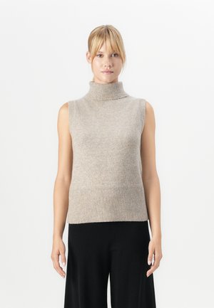 Donna bionda che indossa un maglione senza maniche beige con collo alto e pantaloni neri a gamba larga, in piedi contro uno sfondo bianco semplice.