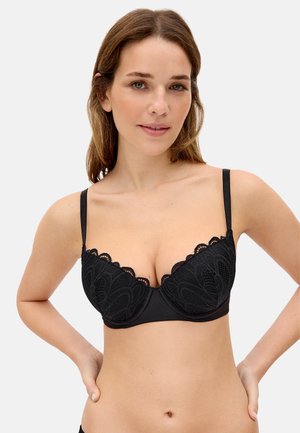 Mujer de piel clara y cabello castaño modelando un sujetador de encaje negro, de pie con las manos en las caderas sobre un fondo blanco liso.