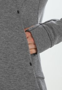 Sudadera gris con un tejido texturizado, que presenta un bolsillo lateral y detalles en negro. Primer plano de una mano metiéndose en el bolsillo.