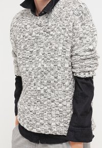 Chunky stickad tröja i ett svartvitt strukturerat mönster, med rundad halsringning och rullade ärmar som är lagda över en svart skjorta.