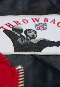 Roter und grauer Stoffpatch mit dem Text "THROWBACK", dem NFL-Logo und einer Grafik von zwei in Bewegung befindlichen Figuren, umrandet in Schwarz und Weiß.
