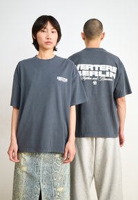 Gray oversized t-shirt made of cotton, featuring white printed text on the front and back. Model wearing light gray sweatpants and a patterned skirt.