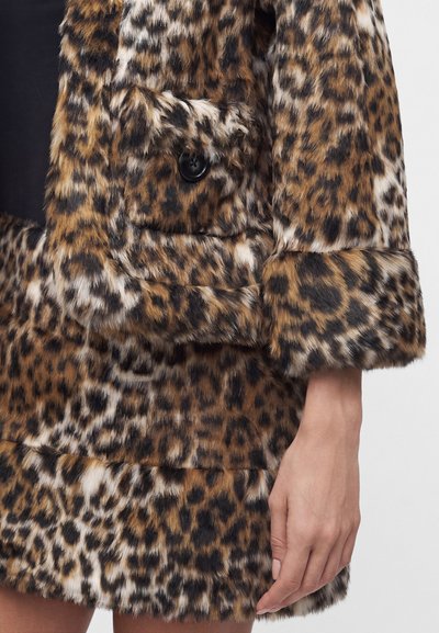 Manteau en fausse fourrure imprimé léopard avec détail de bouton noir sur la manche, porté par une personne avec la main nue visible sur le côté.