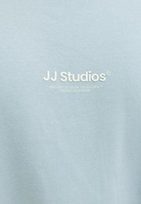 Tricou din bumbac albastru deschis, cu un logo alb "JJ Studios®" și text mai mic care detaliază marca și originea designului.