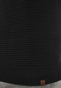 Schwarzer Strickpullover mit geripptem Muster und einem kleinen Leder-Logoetikett am Saum. Der untere Bund hat ein dickes Rippdesign.