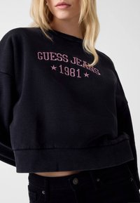 Czarna krótka bluza z prążkowanymi mankietami i dołem, z różowym haftowanym napisem "GUESS JEANS 1981" i gwiazdkami na przodzie.