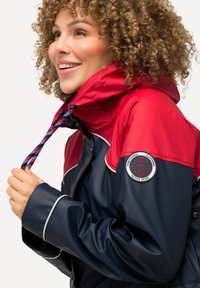Veste imperméable bleu marine et rouge avec une capuche resserrée, un cordon tressé et un patch logo rond sur la manche. Texture lisse et design ajusté.
