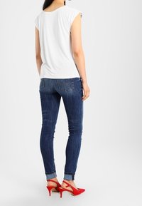 Top blanco de manga corta, jeans ajustados azules con detalles de desvanecimiento y tacones altos rojos con lazos en la parte de atrás.