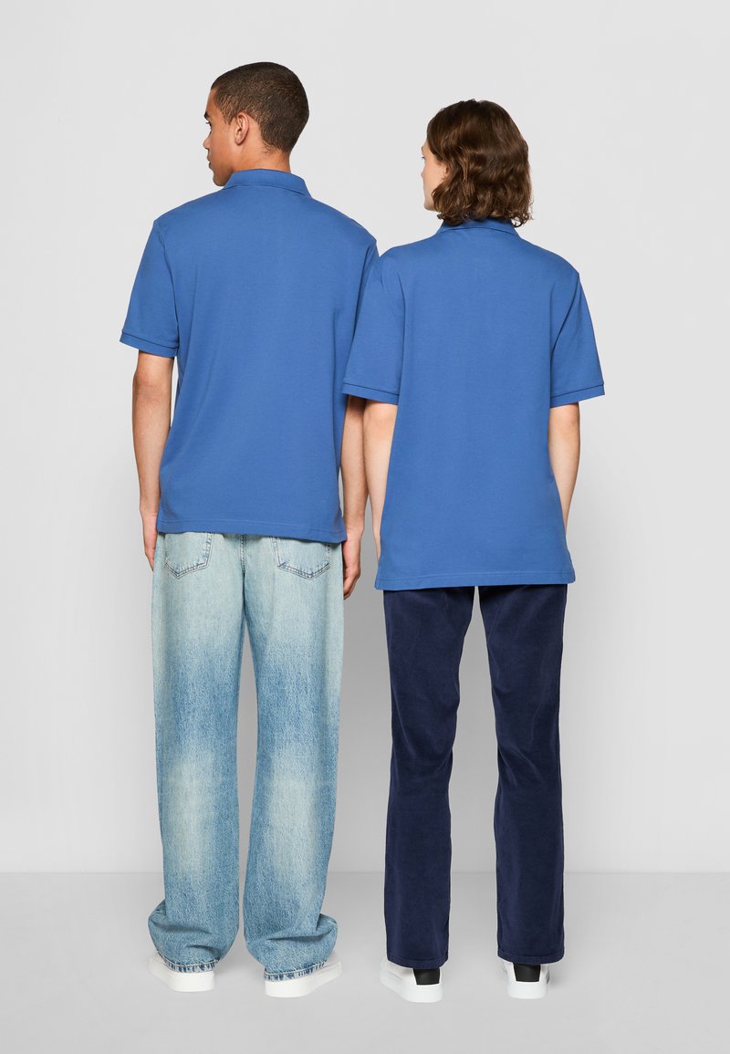 Deux personnes portant des polos bleus à manches courtes, se tenant dos à dos. L'un porte un jean bleu délavé, l'autre un pantalon marine foncé.