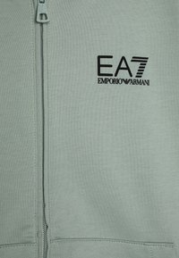 Sweat-shirt zippé vert clair avec une texture lisse. Présente un logo noir "EA7 Emporio Armani" à l'avant.