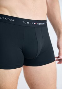 Männlicher Oberkörper trägt schwarze Tommy Hilfiger Boxershorts mit Logo auf dem Hosenbund, dargestellt von der Mitte des Oberschenkels bis zur Taille.