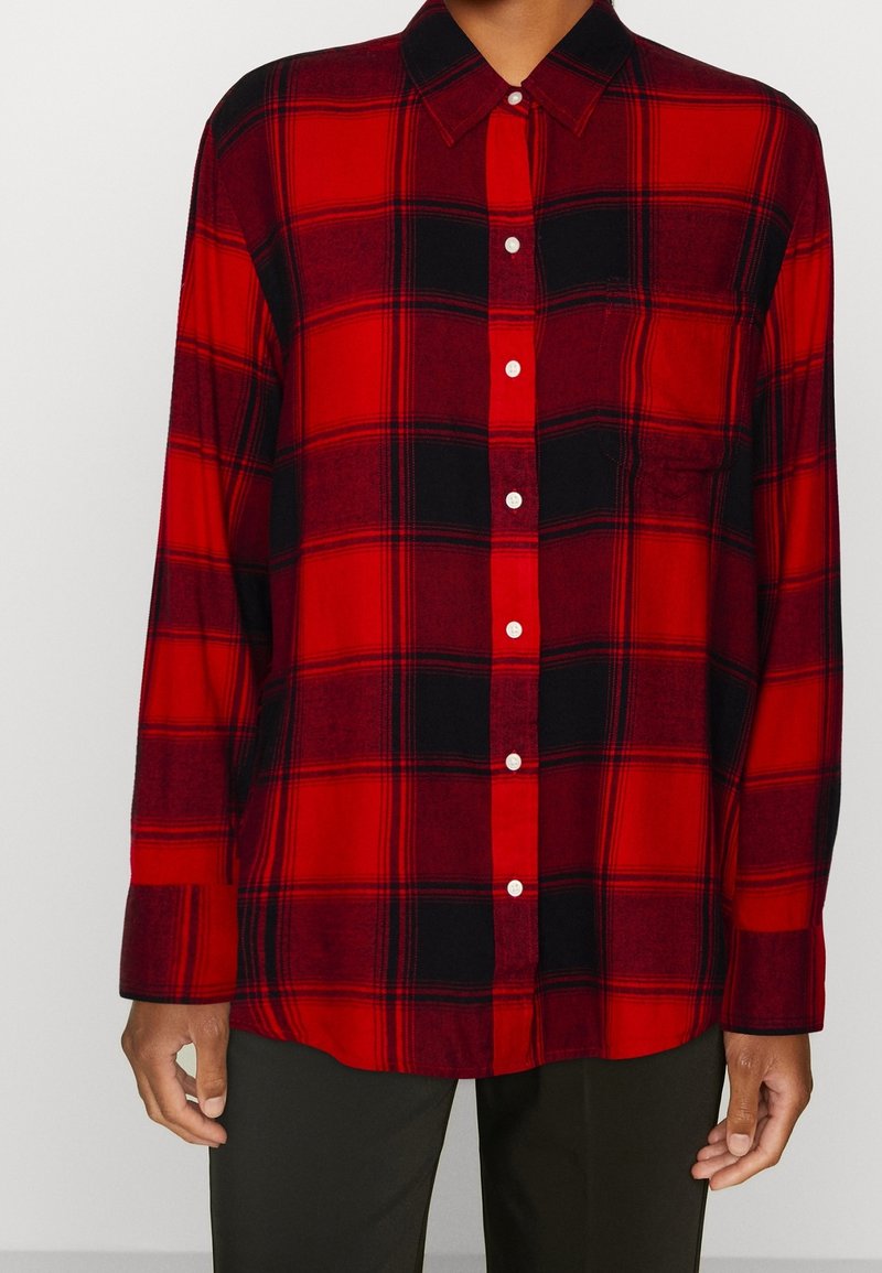 Chemise à carreaux rouge et noir à boutons, manches longues, col et poche avant. Fabriquée en tissu doux avec des coutures visibles.