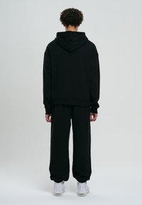 Giovane uomo in piedi di spalle, indossa un felpa nera con cappuccio, pantaloni della tuta neri e scarpe bianche su uno sfondo grigio uniforme.