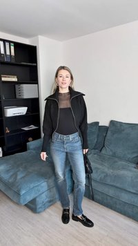 Černá žebrovaná bunda na zip přes průhledný černý top, doplněná světlými modrými skinny džínami a černými botami. Na pozadí světle barevné.