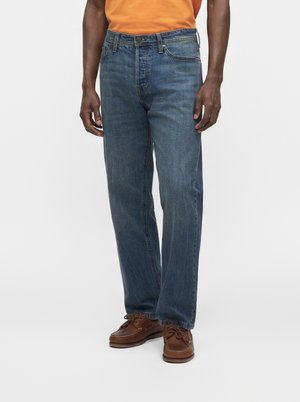 Mann trägt blaue Jeans mit geradem Bein, braune Schnürschuhe und ein oranges Hemd, sichtbar an Taille und Armen.