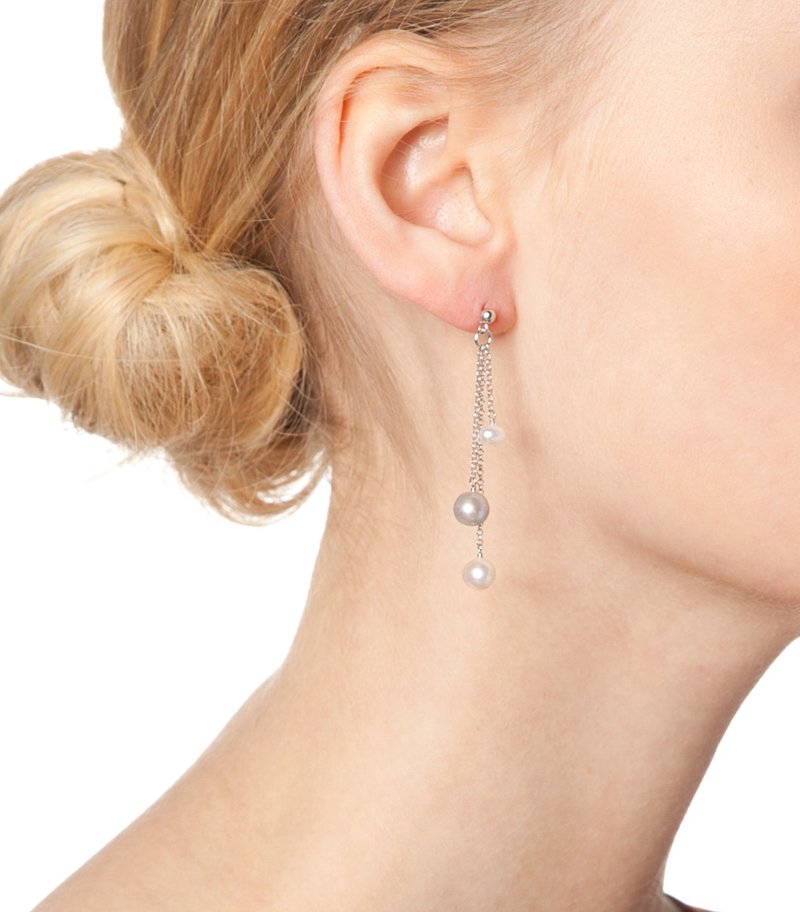 Ezüst lánc fülbevalók, melyeken három gyöngy lóg – egy szürke és két fehér –, finom textúrával és fényléssel, halvány bőrszín mellett.