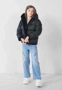 Chaqueta pluma negra con cuello de pelo y cierre de cremallera, combinada con pantalones cargo de pierna ancha en azul claro y zapatillas blancas.