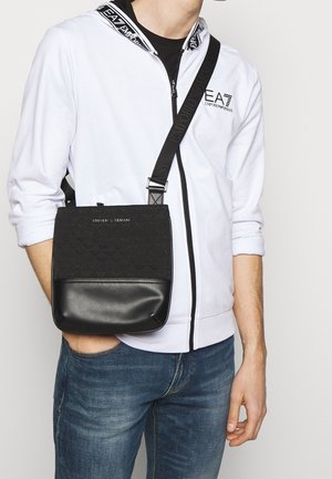 Schwarze Umhängetasche mit einer strukturierten Oberseite mit Logo-Muster und einem glatten unteren Bereich, ergänzt durch einen verstellbaren Riemen.