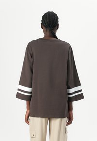 Dunkelbraunes T-Shirt mit Dreiviertelärmeln, das weiße Streifen an den Manschetten aufweist. Der Stoff wirkt weich und hat eine lockere Passform.