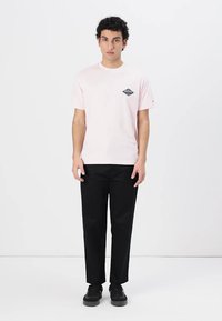 Camiseta de algodón rosa claro con un logo de diamante negro en el pecho, combinada con pantalones negros y zapatos negros.
