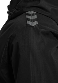 Gros plan de l'épaule d'une veste noire avec un tissu texturé et une bande à motif chevron réfléchissant près d'une capuche noire.