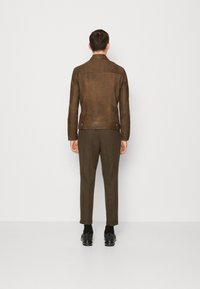Hnědá kožená bunda s přiléhavým střihem, viditelným šitím a zipy. Olivově zelené kalhoty a černé tenisky dotvářejí outfit.