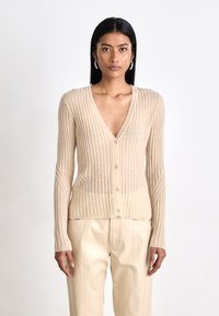 Cardigan beige a coste con scollatura a V, chiusura a bottoni e maniche lunghe. Tessuto morbido, con un design leggermente cropped e orlo grezzo.