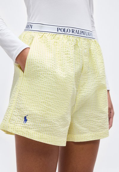 Άτομο που φοράει κίτρινα σορτς από σερσάκερ με μπλε λογότυπο Polo Ralph Lauren και λευκή ελαστική μέση, χέρι στην τσέπη, λευκό μακρυμάνικο πάνω μέρος.