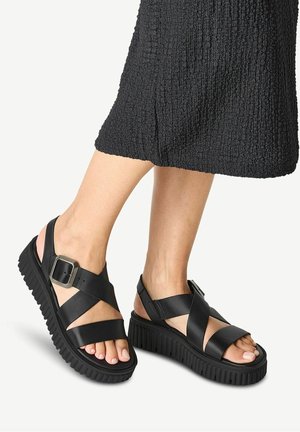 Zwarte platformsandalen met brede gekruiste banden, voorzien van een gespsluiting aan de zijkant, een dikke gestructureerde zool en een open achterkant.