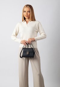 Schwarze Handtasche aus Leder mit goldenen Beschlägen, strukturiertem Finish, oberen Griff und abnehmbarem Gurt, gehalten von einer Person in einem hellen Sweater und beigen Hosen.