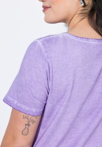 Camiseta de color púrpura claro con mangas cortas, escote redondo y textura suave. Presenta un tatuaje visible en el brazo.