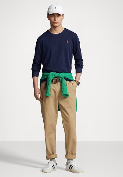 Jūras spēku garām piedurknēm krekls, zaļš džemperis, kas sasiets ap vidu, khaki bikses un baltie sporta apavi ar zilām svītrām; krekla priekšpusē ir polo logotips.