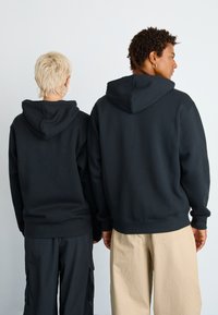 Des sweats à capuche noirs à coupe décontractée avec une poche kangourou et des poignets côtelés, portés par deux personnes tournées le dos. L'un porte un pantalon noir, l'autre un beige.