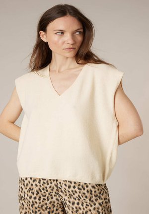 Femme aux cheveux bruns portant un pull sans manches crème à col en V et un pantalon imprimé léopard, regardant sur le côté avec les mains derrière le dos.