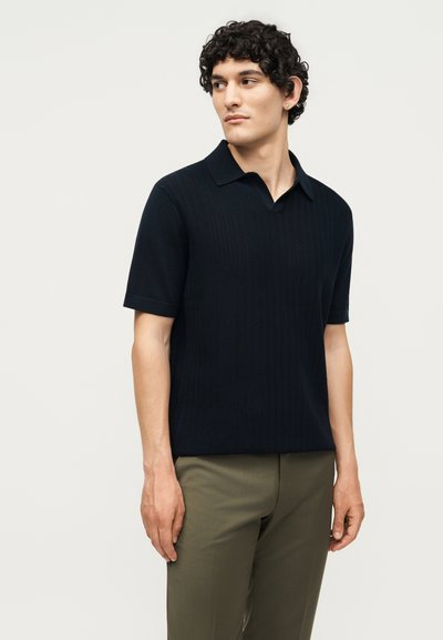 Νέος άνδρας με κατσαρά μαλλιά που φοράει μαύρο πλεκτό πόλο με κοντά μανίκια και χακί παντελόνια, κοιτάζοντας στο πλάι μπροστά σε απλό φόντο.