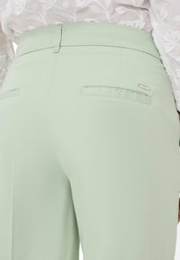 Lichtgroene broek van gladde stof, met een hoge tailleband, twee achterzakken en een klein accent van zilverkleurig materiaal.