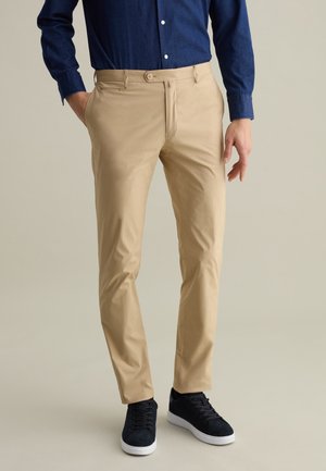 Beige Chinos mit Slim-Fit und glatter Textur, ausgestattet mit einem Knopfverschluss vorne und Seitentaschen, kombiniert mit schwarzen Sneakers.