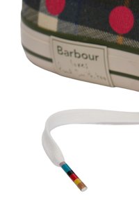 Τακούνι παπουτσιού με ετικέτα «Barbour» σε καρό ύφασμα και λευκό κορδόνι με χρωματιστές ρίγες στην άκρη, πάνω σε λευκό φόντο.